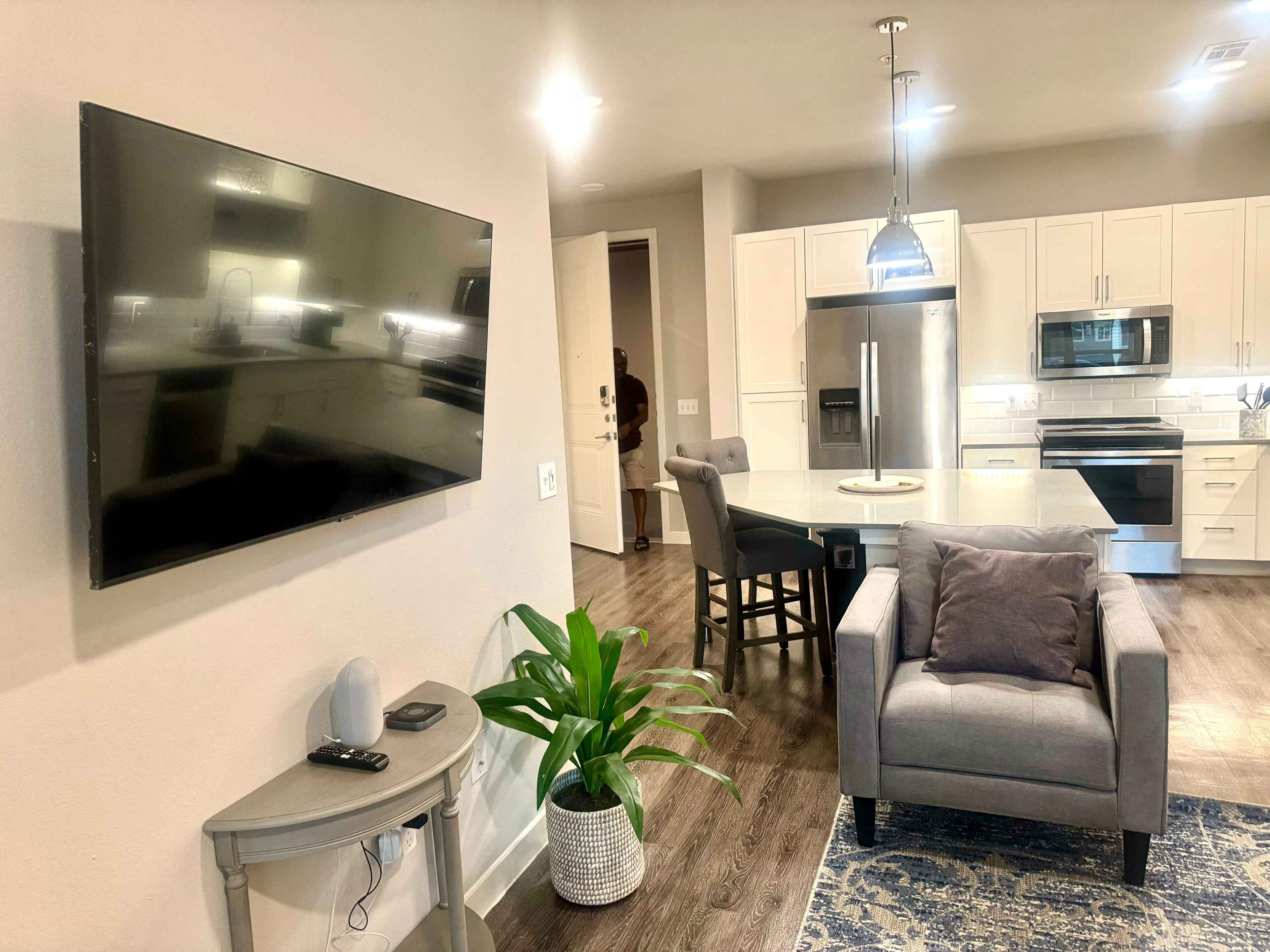 Una cocina moderna con electrodomésticos de acero inoxidable, un comedor con mesa y sillas, un sofá gris y un televisor de pantalla plana montado en la pared.