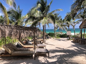 Un salon de plage doté de mobilier en bois, de palmiers et d’une structure à toit de chaume.