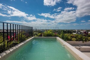 A rooftop pool with a clear view of the cityscape, surrounded by lush greenery and a metal railing.