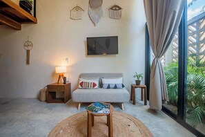 A cozy room with a bed, a bedside table with a lamp, a small table with a magazine, and a large window with curtains. - Bright & Sunny 2BR Loft w/ Roof Pool in Downtown (Tulum)