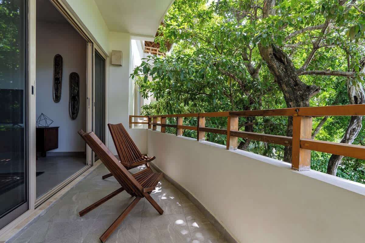 A balcony with two wooden chairs, a glass door leading inside, and a view of lush green trees.