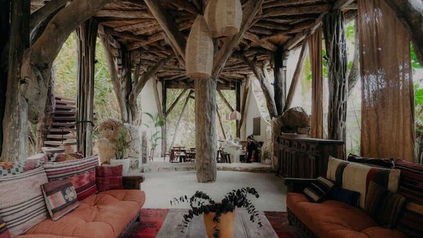 Un interior rústico con vigas de madera, una zona de estar central con cojines y un jarrón grande con follaje negro.