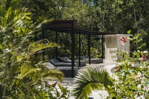 Uma área de estar coberta ao ar livre, com espreguiçadeiras e uma pérgola de madeira, rodeada por vegetação exuberante.