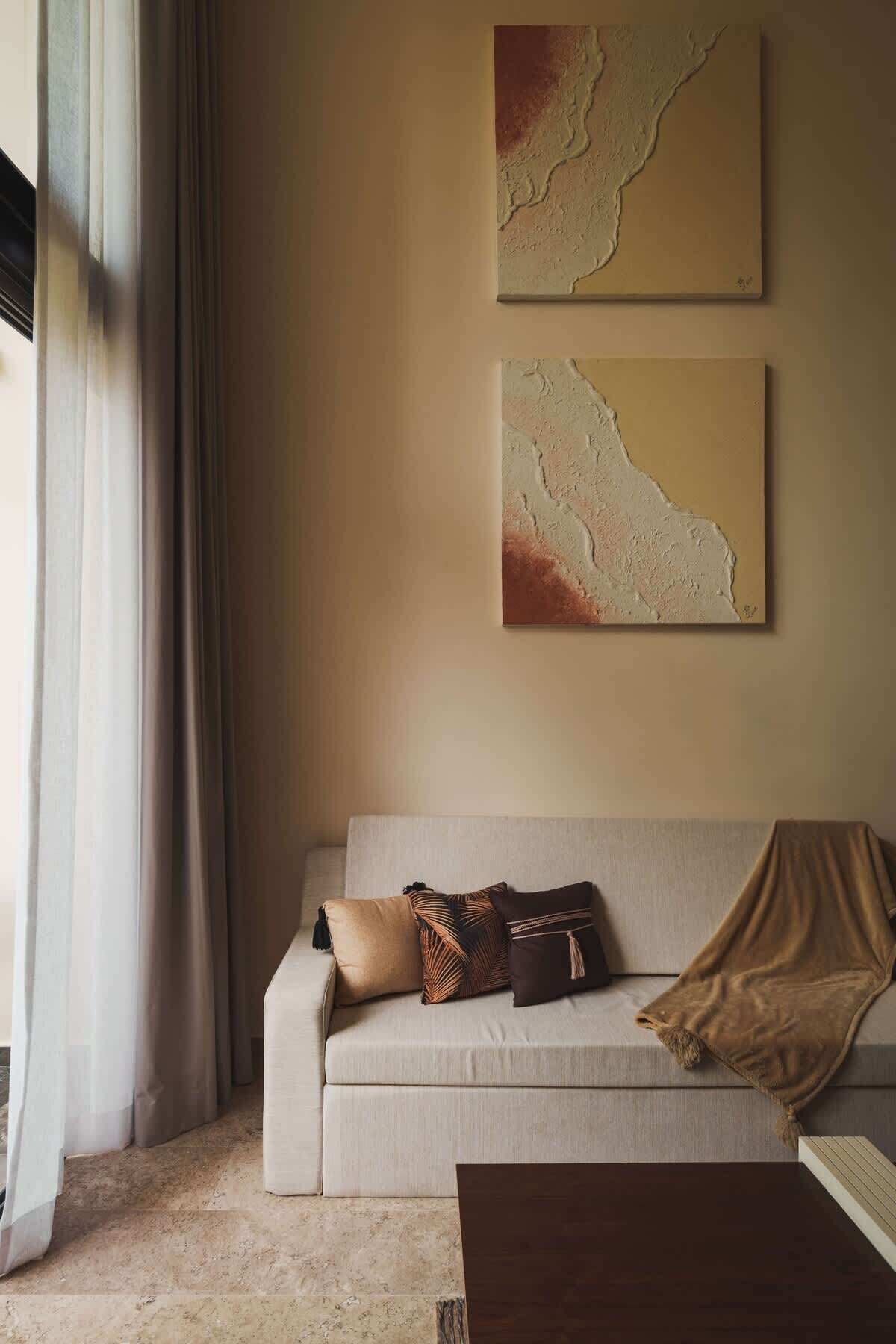 Ein Wohnzimmer mit einem beigefarbenen Sofa, zwei abstrakten Wandbildern und einem fenster mit Vorhängen.