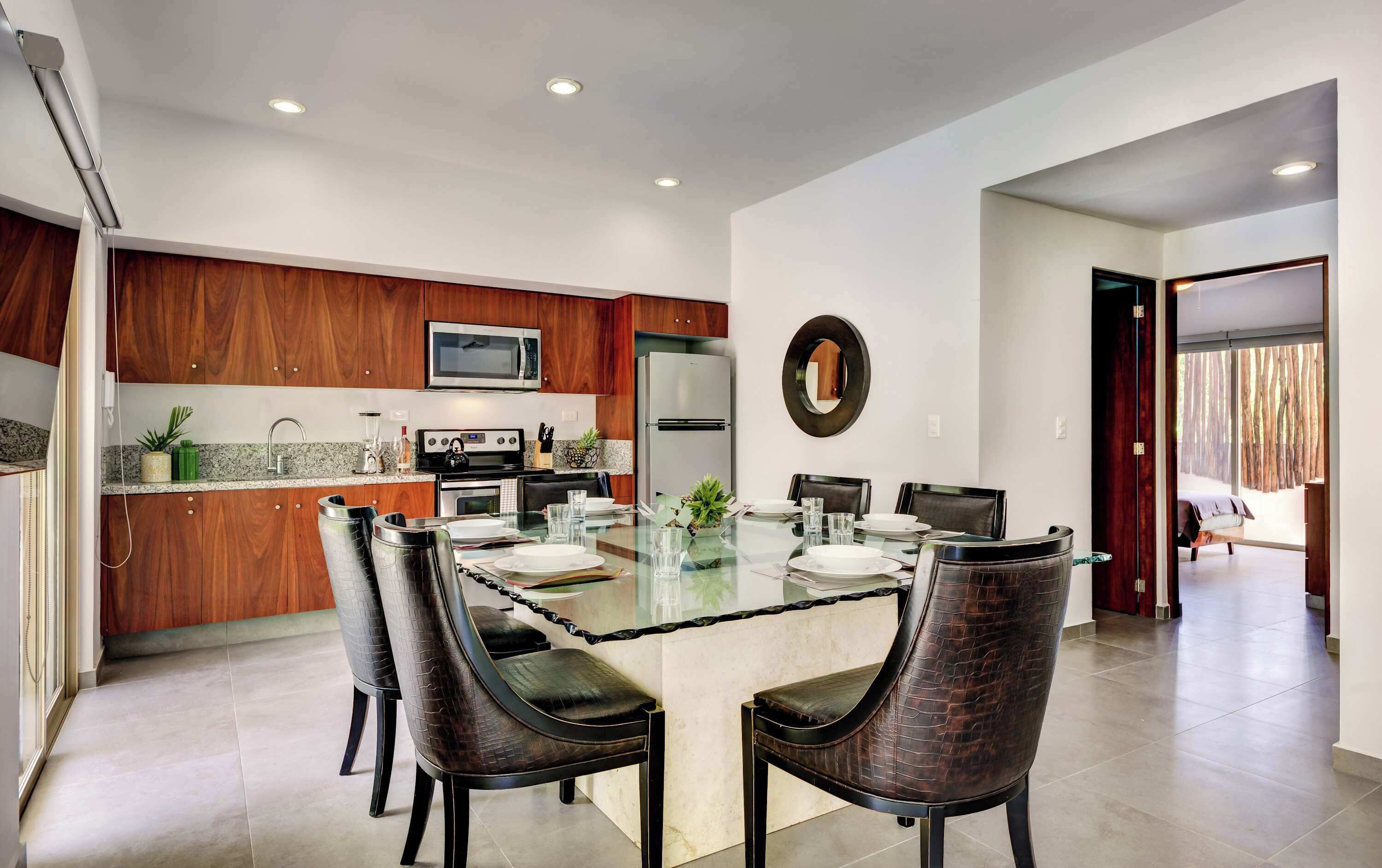 A modern kitchen with wood cabinets, a central island, and a dining area with a glass table and chairs.