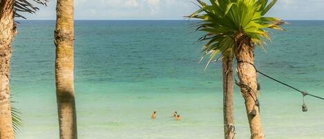 A beach with palm trees, clear turquoise water, and people swimming and relaxing on the sand.