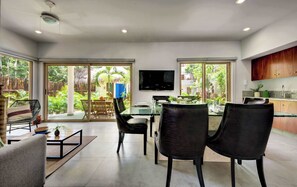 A modern dining area with a glass table and chairs, a flat-screen TV mounted on the wall, and a view of a lush garden through large sliding doors.