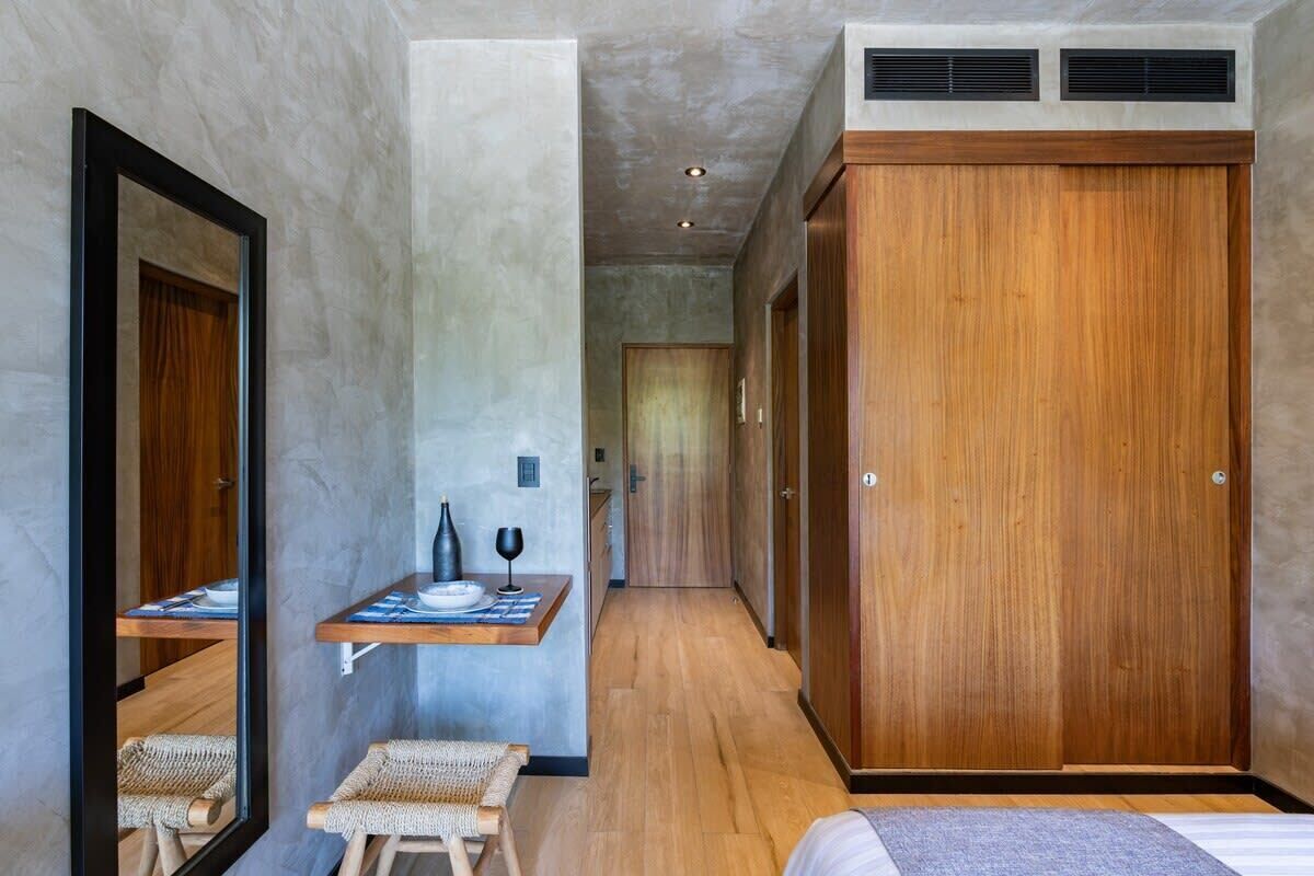 Una habitación de hotel moderna con un espejo grande, una pequeña mesa de comedor con un florero y una copa de vino, un armario de madera y una cama con un tapete gris.