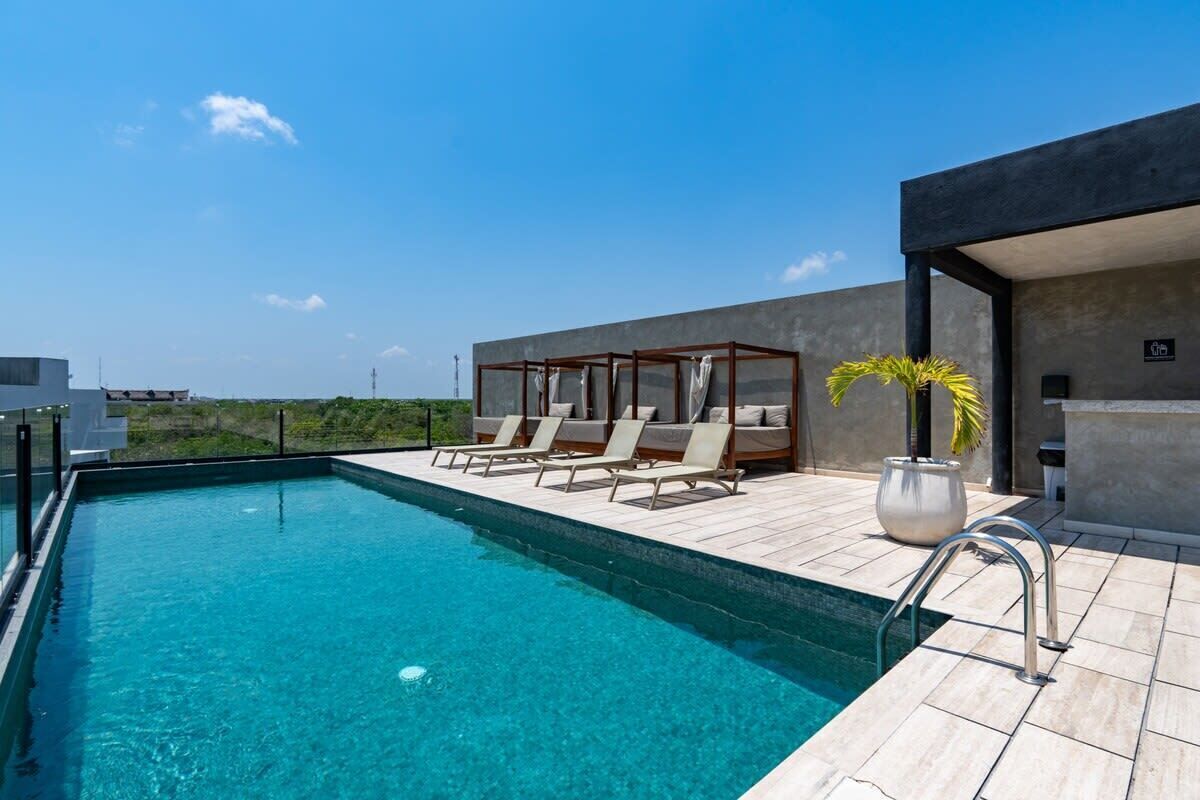 Une piscine sur le toit avec une eau d’un bleu cristallin, des chaises longues, un espace salon couvert et un bâtiment moderne offrant une vue sur le paysage.