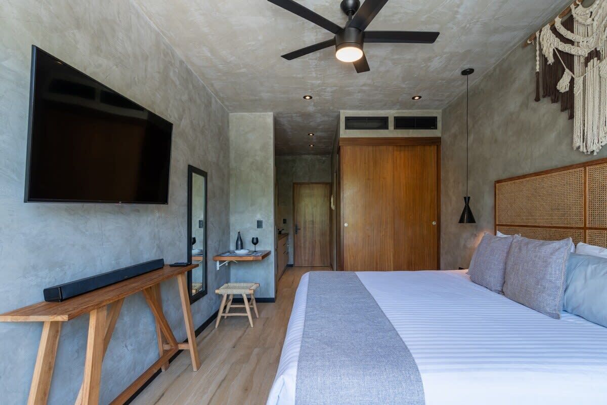 A modern bedroom with a large bed, a ceiling fan, a flat-screen TV mounted on the wall, and a wooden nightstand.