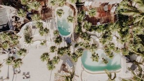 Vue aérienne d’un complexe hôtelier doté de plusieurs piscines, entouré de palmiers et bordé d’une plage de sable.