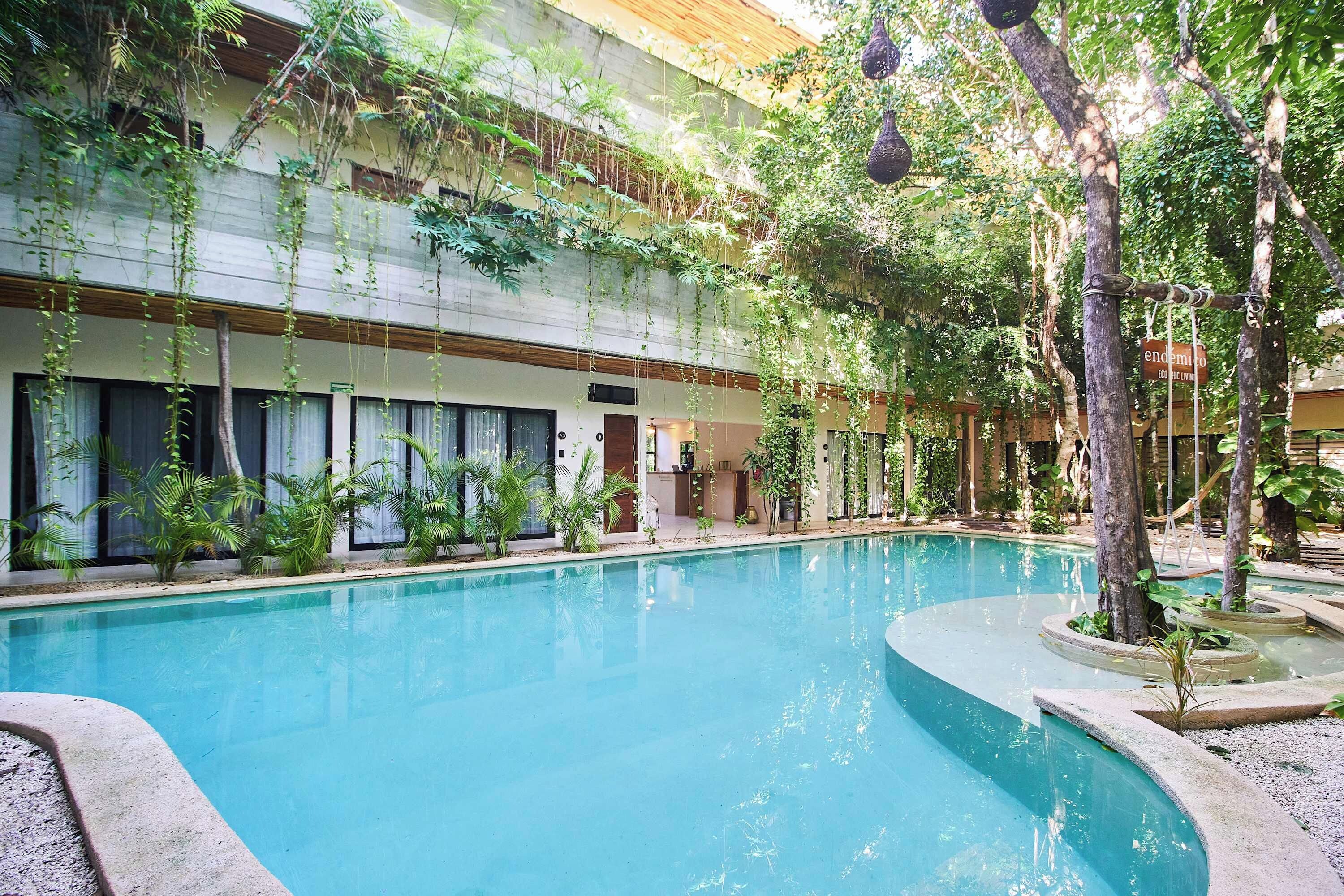 A pool area with a clear blue pool, surrounded by greenery and a building with windows.