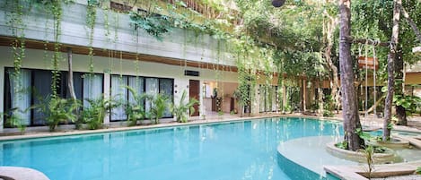 Un espace piscine avec une eau d’un bleu limpide, entouré de verdure et bordé d’un bâtiment doté de fenêtres.