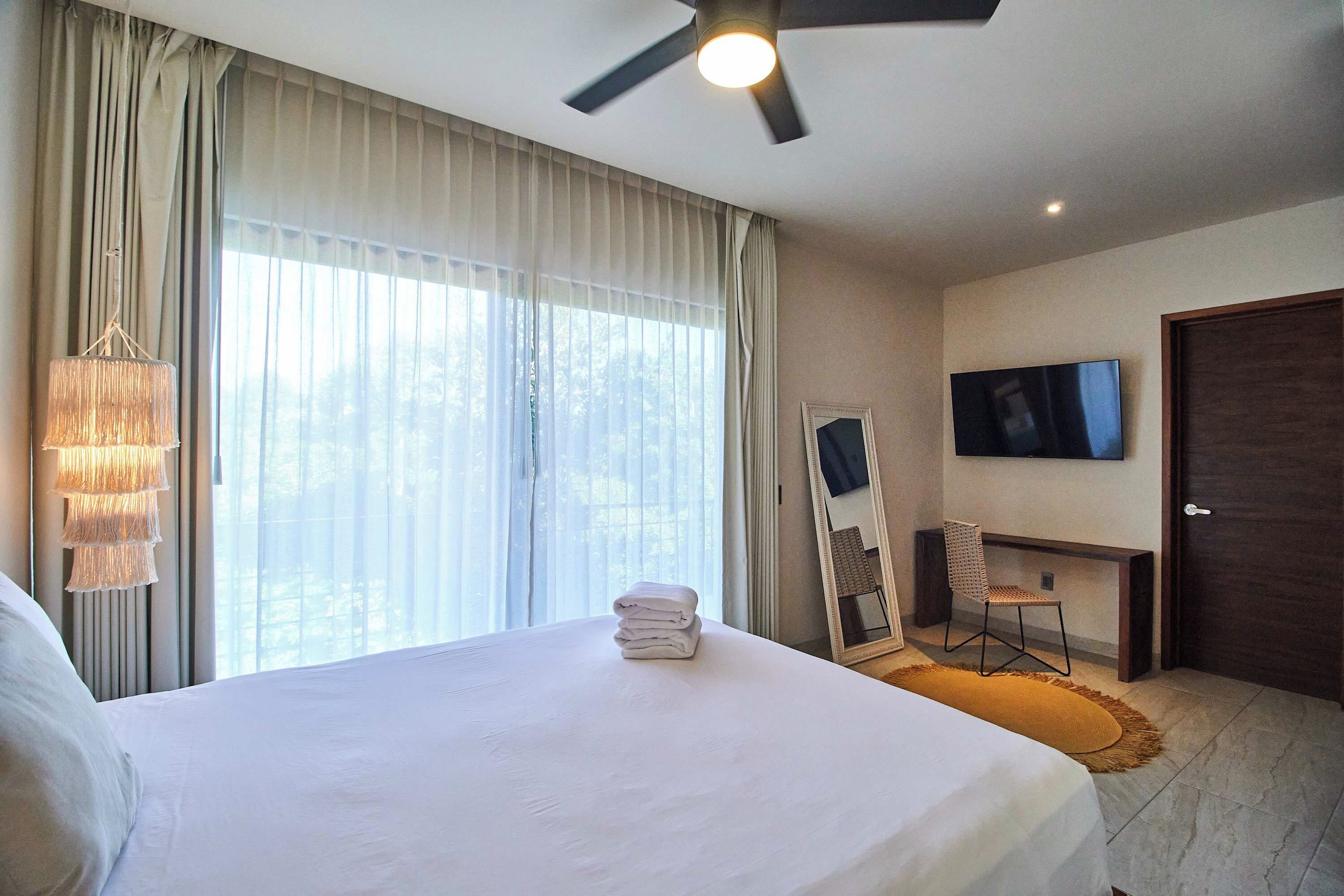 Una habitación de hotel con cama, televisor montado en la pared, ventilador de techo y ventana con cortinas.