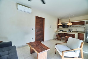 A modern living room with a sofa, a wooden coffee table, and a grey armchair. A kitchen with wooden cabinets and a built-in island is visible in the background.