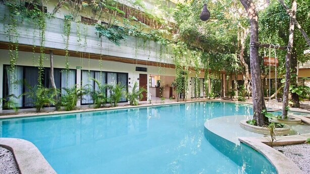 An indoor swimming pool with a curved edge, surrounded by greenery and a building with large windows. - Tulum Penthouse with Rooftop Pool and BBQ Grill (Tulum)