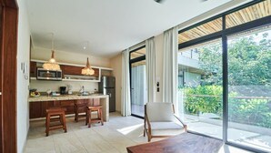 A modern kitchen with wooden cabinets, a dining area with a wooden table, and a large sliding glass door leading to a balcony with greenery. - Tulum Penthouse with Rooftop Pool and BBQ Grill (Tulum)