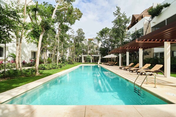 A swimming pool with lounge chairs and a pergola, surrounded by greenery and a building.