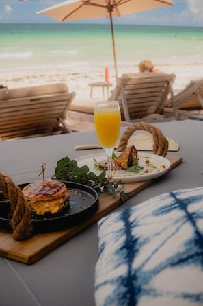 A beachside dining setup with a plate of food, a glass of orange juice, and a wooden table. - Premium Condo with 2 Pools, Gym, Yoga Deck & BBQ (Tulum)