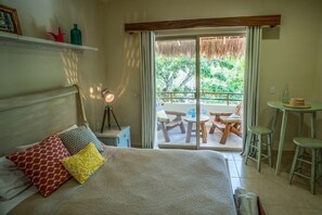 Un dormitorio con cama, mesita de noche, lámpara, ventana con vista a un balcón y una pequeña mesa con sillas.