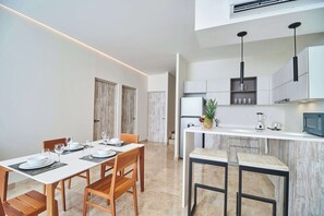 Une cuisine moderne avec un îlot central, un coin repas avec une table et des chaises blanches, et un couloir menant à une porte.