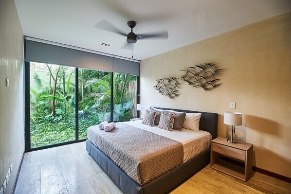 Un dormitorio moderno con cama, mesita de noche, ventilador de techo y vistas al verdor a través de amplios ventanales.
