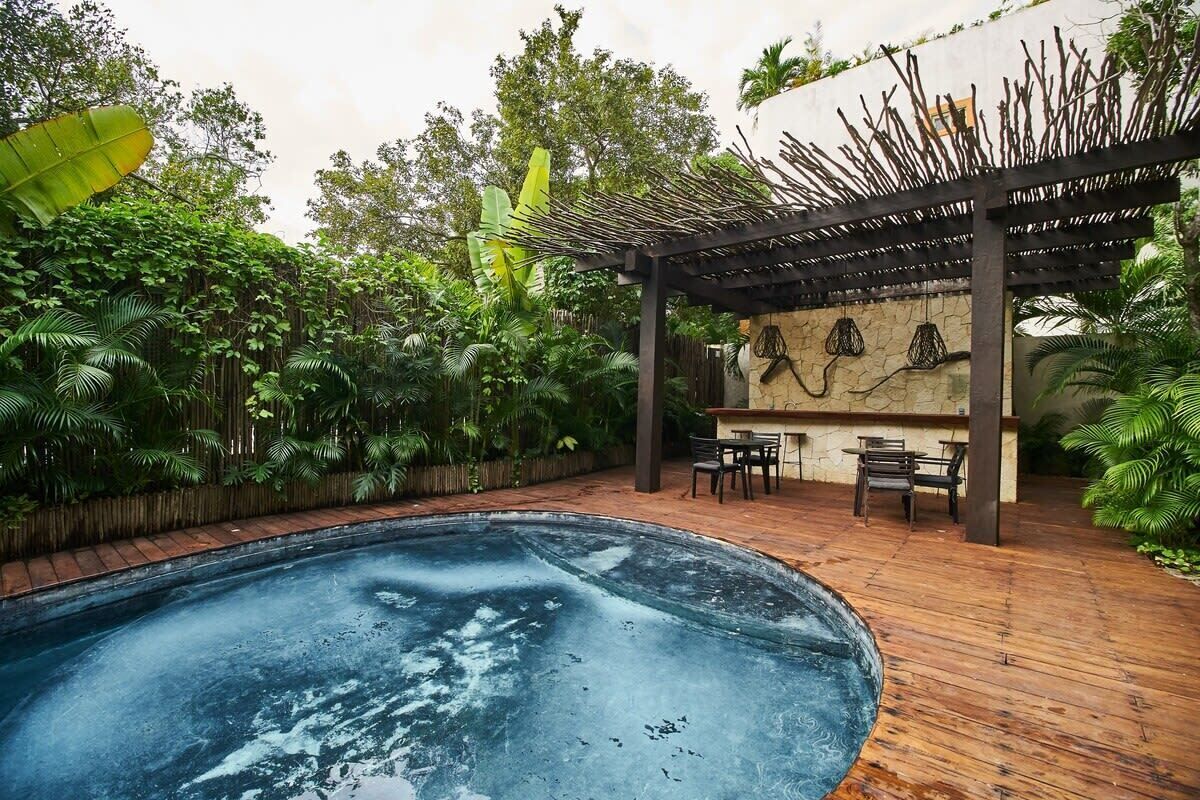 A swimming pool surrounded by lush greenery, a wooden deck, and a pergola with hanging lights.