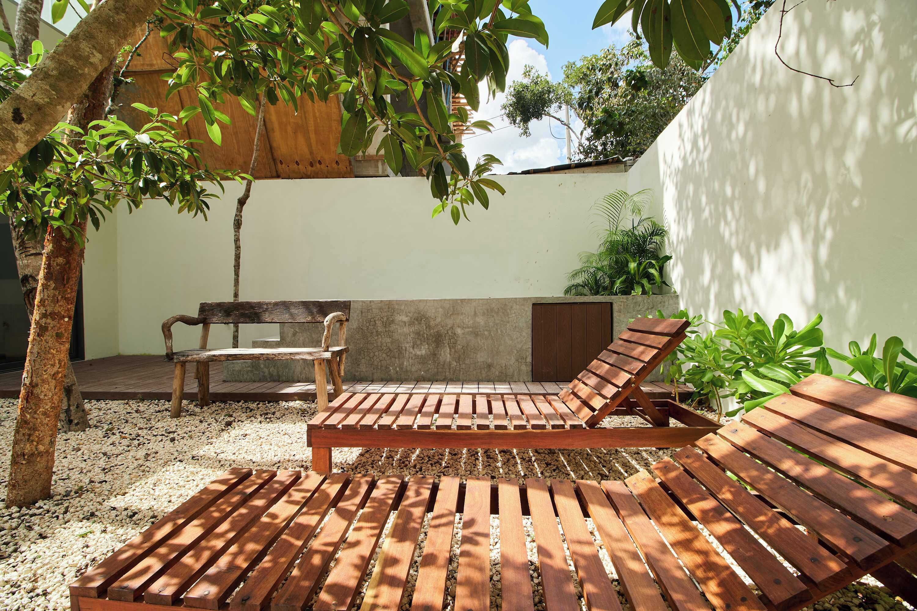 A wooden bench and a lounge chair in a shaded outdoor area with a concrete wall and potted plants.