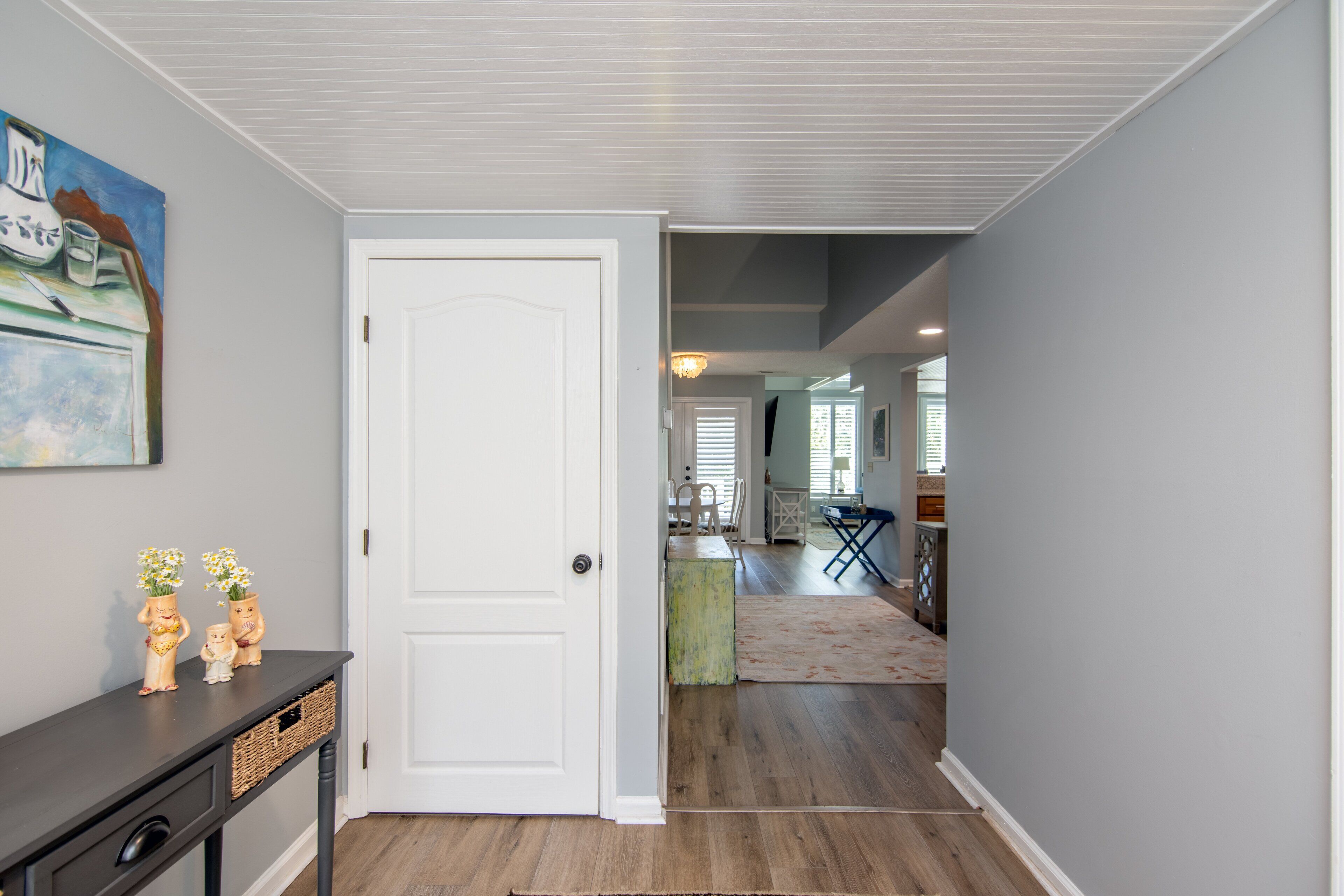 A hallway with a white door, a dark console table with decorative items, and a painting on the wall.