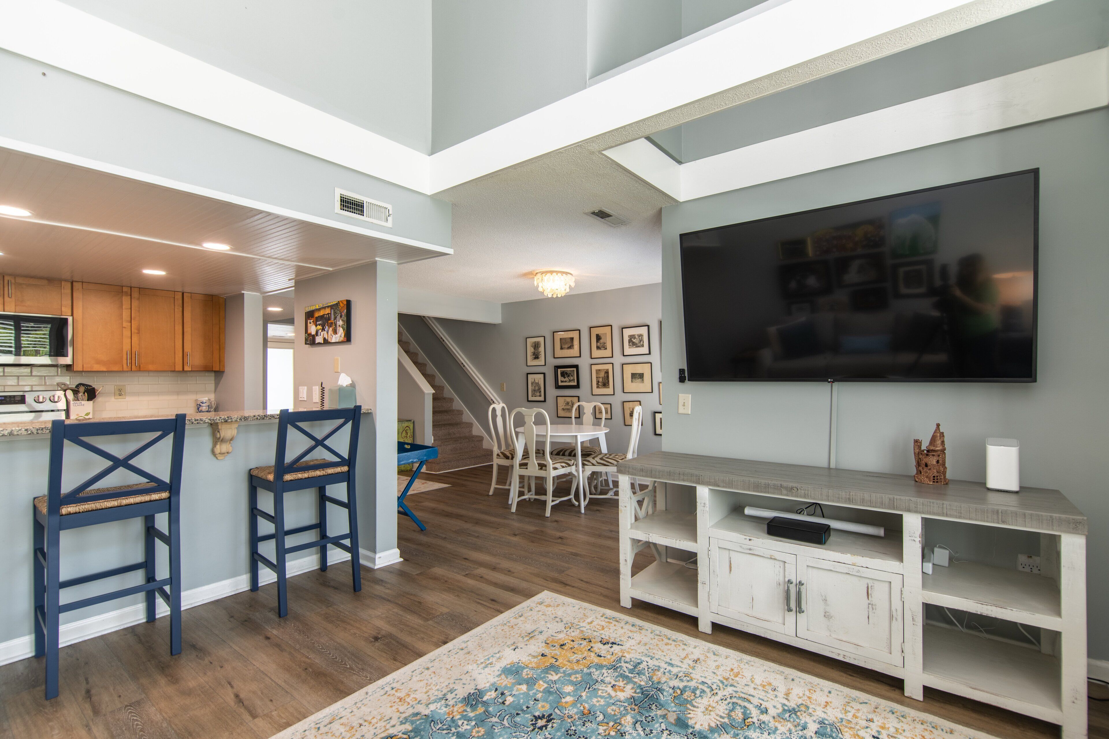 A modern kitchen with wooden cabinets, a bar area with blue chairs, a dining table with chairs, and a large flat-screen TV mounted on the wall.