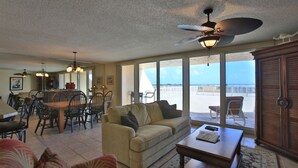 A spacious living room with a large window overlooking the beach, a dining area with a table and chairs, and a ceiling fan.