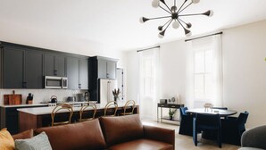 Eine moderne Küche mit dunklen Schränken, einer Mikrowelle und einem Kühlschrank. Ein Essbereich mit einem runden Tisch und Stühlen ist vorhanden. Im Wohnzimmer befindet sich ein braunes Ledersofa.