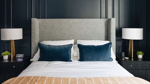 Ein ordentlich bezogenes Bett mit grauem Kopfteil, weißer Bettwäsche und zwei blauen Kissen. Zwei Nachttische mit Lampen und kleinen Topfpflanzen sind vorhanden.