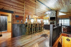 Ein rustikales Blockhaus-Interieur mit Holzwänden, einem Kamin aus Stein, einer Bar mit Hockern und einem an der Wand befestigten Fernseher.