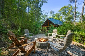 Une terrasse en bois, dotée de chaises Adirondack et d’un foyer, est entourée d’une forêt.