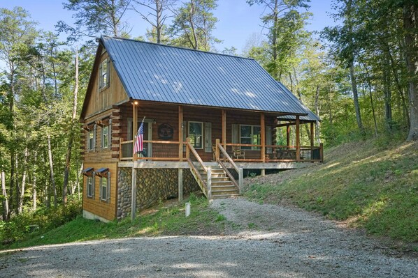 A rustic cabin with a stone foundation, wooden exterior, and a metal roof. It features a porch with a staircase leading to a deck. - Freedom Ridge Cabin | Hocking Hills, Hot Tub (Logan)