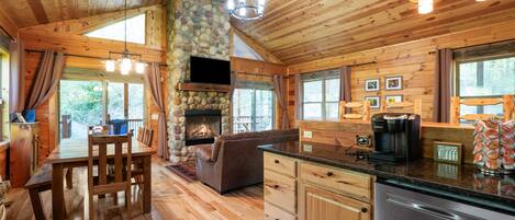 L’intérieur d’un chalet rustique est agrémenté d’une cheminée en pierre, d’une table à manger en bois et d’un coin cuisine équipé d’une machine à café.
