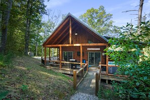 A wooden cabin with a covered deck, a hot tub, and a large window overlooking the forest. - Freedom Ridge Cabin | Hocking Hills, Hot Tub (Logan)