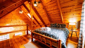 A wooden cabin bedroom with a bed, a small table, a lamp, and a ceiling fan.