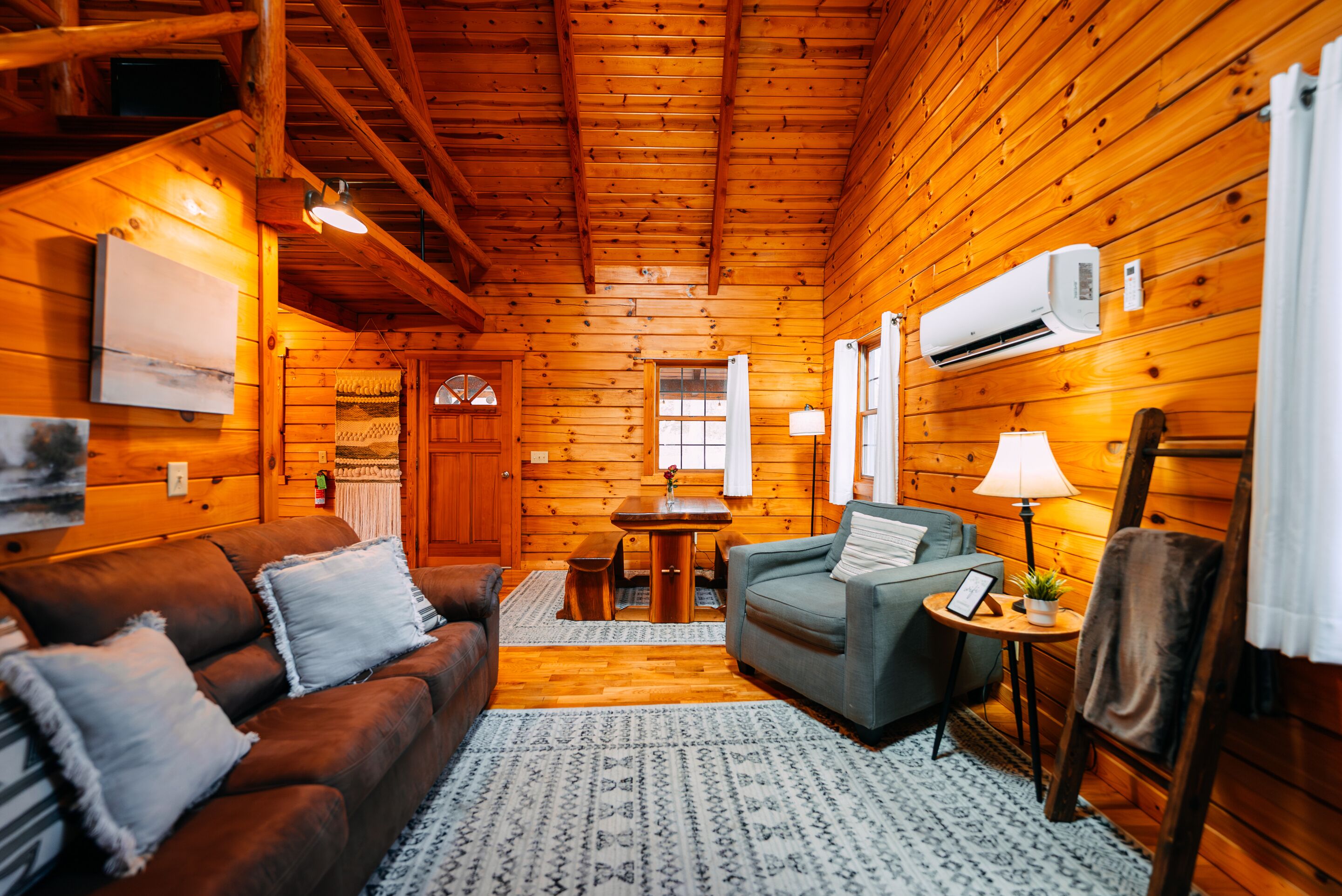 A cozy cabin interior with a wooden ceiling, a sofa, a chair, a small table, and an air conditioning unit.