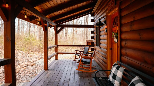 A wooden cabin with a porch, rocking chairs, a bench, and a view of the forest.