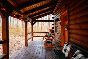 Una cabaña de madera con un porche, mecedoras, un banco y vistas al bosque.