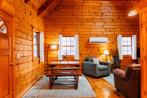 Interior de una cabaña de madera con paredes de tablones, una mesa de comedor, un sofá y un equipo de aire acondicionado.
