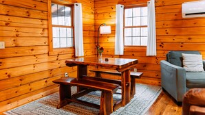 A wooden cabin interior with a wooden table, benches, a sofa, and two windows with curtains.