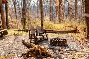 Ein bewaldeter Campingplatz mit zwei Adirondack-Stühlen, einer Feuerstelle und einer Holzbank.