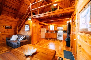 Interior de una cabaña de madera con sofá, mesa de comedor, zona de cocina y un altillo.