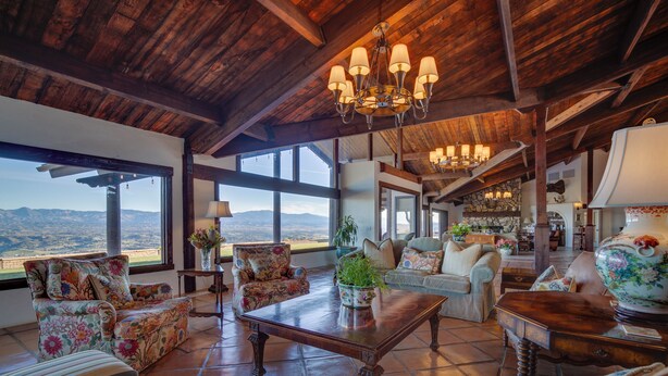 A spacious living area with a high ceiling, large windows offering a view of the mountains, and a variety of seating options including floral-patterned sofas and armchairs.