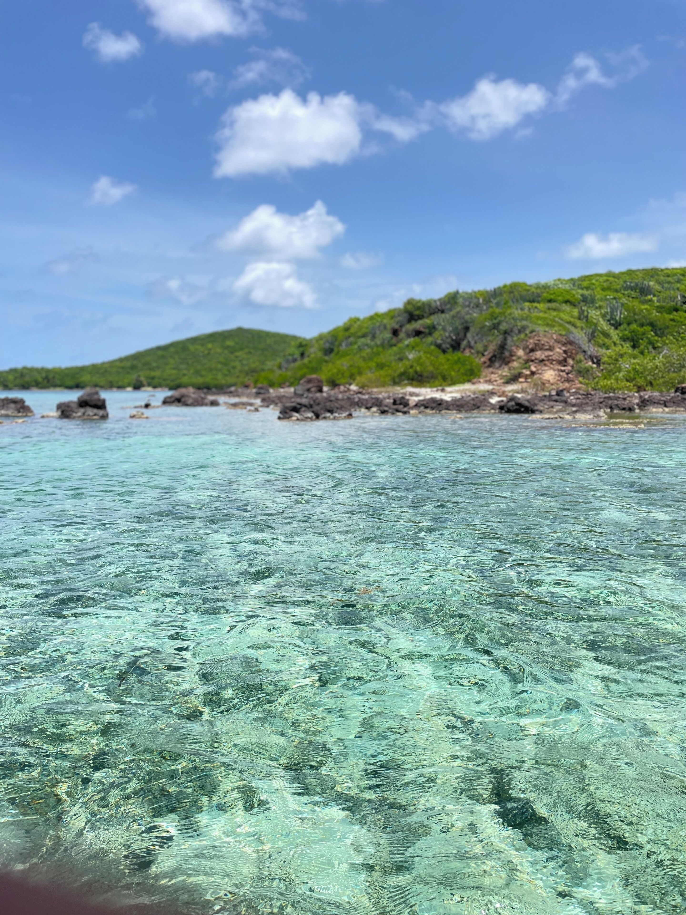 A clear view of a beach with turquoise water, rocky outcrops, and green hills in the background.