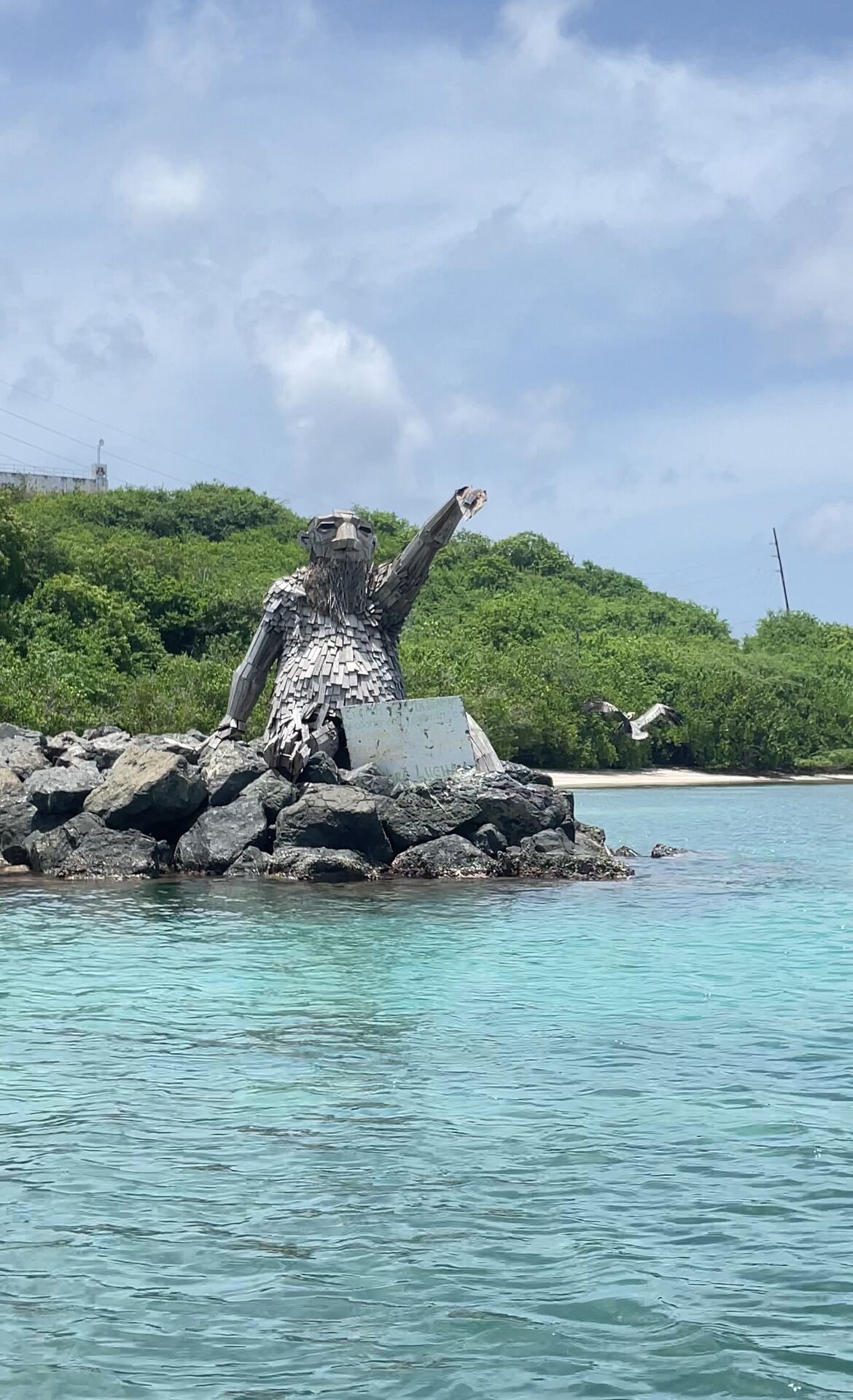 A sculpture on a rocky outcrop surrounded by clear turquoise water and a green hillside.