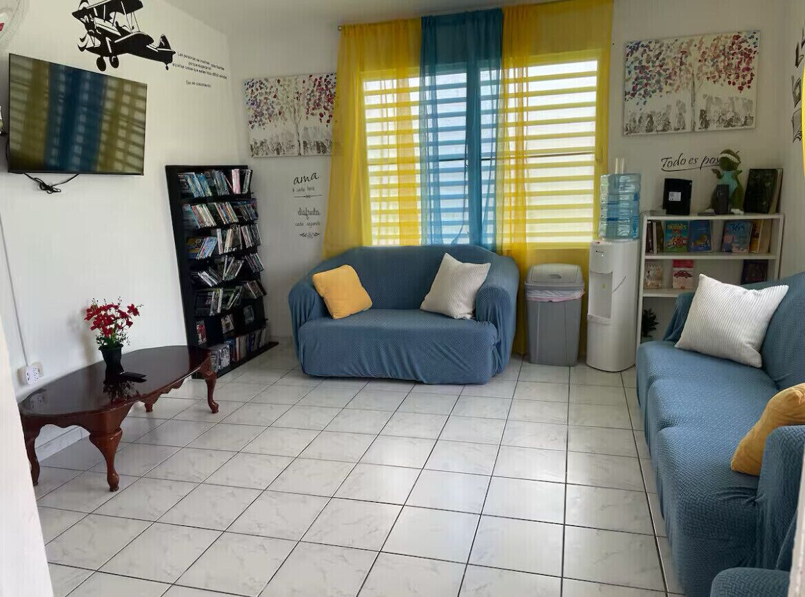 Un salon avec un canapé bleu, une bibliothèque, un téléviseur et une fenêtre ornée de rideaux jaunes.